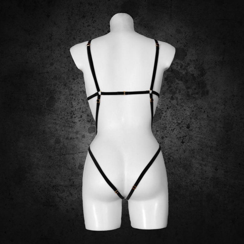 May include: Black lingerie body harness with gold-coloured hardware. The harness features multiple straps that connect at the shoulders, back, and waist. The garment is displayed on a white mannequin against a dark grey background.