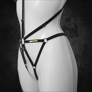 May include: Black lingerie harness with gold-tone metal rings and accents. The harness features multiple straps and a small gold-tone nameplate. The lingerie is displayed on a white mannequin against a dark background.