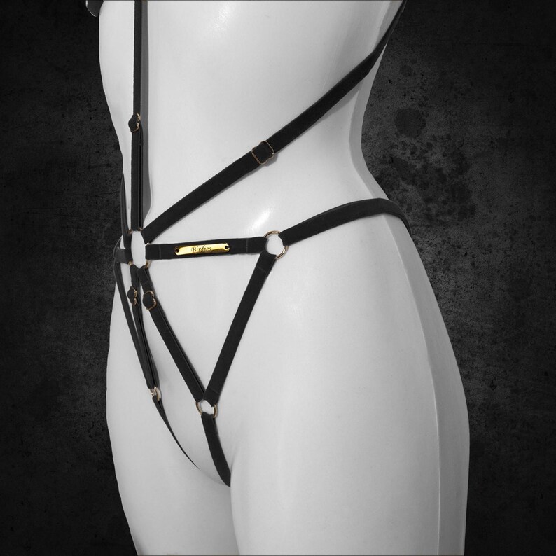 May include: Black lingerie harness with gold-tone metal rings and accents. The harness features multiple straps and a small gold-tone nameplate. The lingerie is displayed on a white mannequin against a dark background.