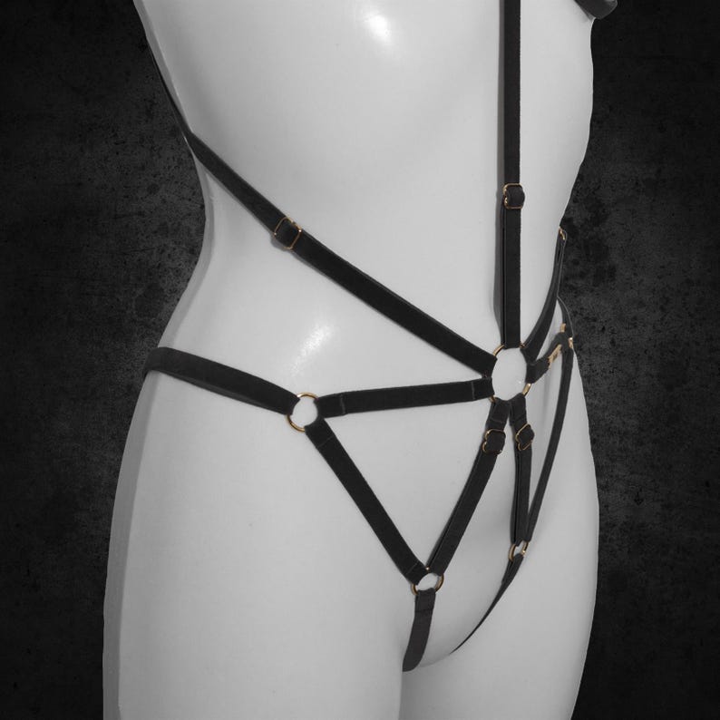 May include: Black lingerie harness with gold-coloured metal rings and buckles. The harness features multiple straps that create a geometric design across the torso and hips. The garment is displayed on a white mannequin.