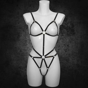May include: Black body harness with adjustable straps and metal ring accents. The lingerie-style harness features a triangle top and a low-rise bottom, displayed on a white mannequin against a dark background.