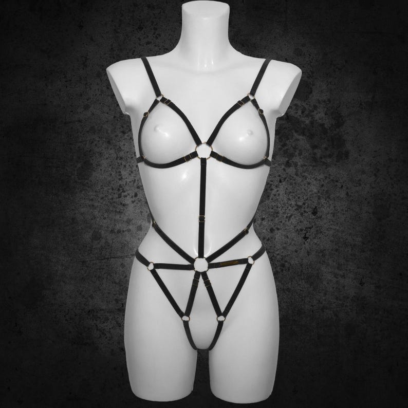 May include: Black body harness with adjustable straps and metal ring accents. The lingerie-style harness features a triangle top and a low-rise bottom, displayed on a white mannequin against a dark background.