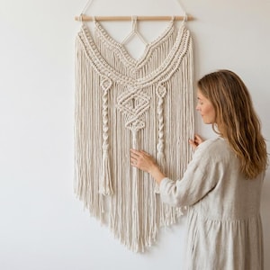 Può includere: Un grande arazzo da parete in macramè color crema, con intricati motivi tessuti e frange. L'opera d'arte tessile è sospesa a un tassello di legno e pende contro un muro bianco. Il design presenta forme geometriche e dettagli annodati.