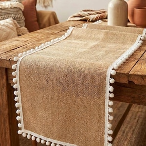 May include: A rectangular burlap table runner with white pom-pom trim. The runner is a natural tan color and is placed on a rustic wooden table. The pom-poms are evenly spaced along the edges.