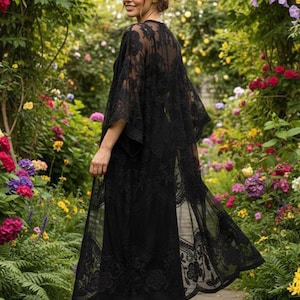 May include: A black lace kimono-style cover-up with wide sleeves and a flowing, open front. The intricate floral lace design is visible against a darker underlayer. The garment is worn in a garden setting with colorful flowers.