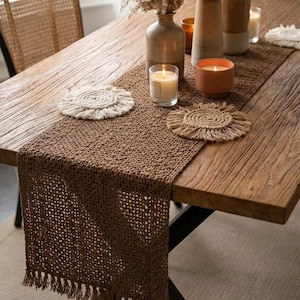 Puede incluir: Una mesa de madera rústica está decorada con un camino de mesa tejido marrón y posavasos a juego. Jarrones decorativos con flores secas y velas encendidas en soportes de vidrio y terracota añaden calidez a la escena. Una silla tejida está en el fondo.