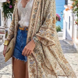 May include: A beige floral print kimono with long sleeves, worn over a white lace camisole and denim shorts. The kimono features a detailed pattern in shades of brown, blue, and green. Accessories include a gold clutch, bracelets, and a belt.