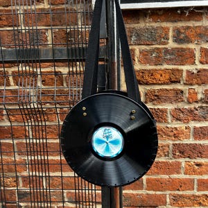 May include: A black vinyl record bag with a black strap. The bag has a blue and white label with a ship design. The bag is suspended against a brick wall and a black metal fence.