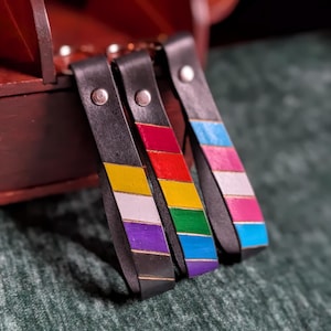 May include: Three black leather keychains featuring painted diagonal stripes. The first displays yellow, white, and purple. The second shows red, yellow, and green. The third features blue, pink, white, pink, and blue. Each has a silver metal rivet.