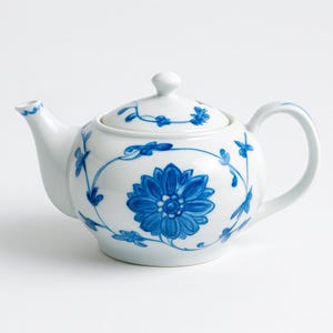 May include: White ceramic teapot with a blue floral design. The teapot has a large blue flower on the front, with smaller blue floral accents and vines. The lid has a small knob and is also decorated with blue floral patterns.