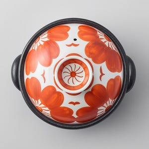 May include: Overhead view of a ceramic cooking pot with a lid. The lid is white with a repeating pattern of orange floral designs. The pot has a black rim and handles.