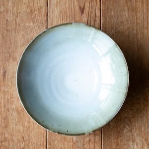Puede incluir: Un cuenco redondo de cerámica azul claro con una superficie ligeramente texturizada. El cuenco tiene un borde ancho con un tono más oscuro y terroso. El cuenco está sobre un fondo de madera.