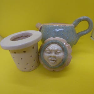 May include: A ceramic tea set with a whimsical design. The set includes a small teapot with a handle, a tea infuser, and a lid with a face design. The teapot is green with blue dots, and the other pieces are cream-coloured.