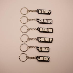 May include: Six keychains with silver rings and chains. Each keychain has a black rectangular tag with a different name in white block letters: HENRY, OLIVER, OSCAR, HARRY, GHEO, and JACK. The keychains are arranged vertically.