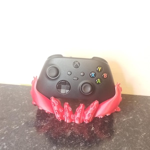 Puede incluir: Un mando de Xbox negro con botones de colores, colocado sobre un soporte rojo con forma de tentáculo. El mando tiene un botón central de Xbox, dos joysticks y un panel direccional. El soporte es de color rojo vivo.
