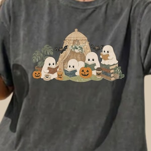 Könnte beinhalten: Ein anthrazitfarbenes T-Shirt mit einem Grafikdruck von vier weißen Geistern, die in und um ein Zelt herum Bücher lesen. Das Design zeigt orangefarbene Kürbisse, grüne Topfpflanzen, schwarze Fledermäuse und einen Stapel Bücher in gedeckten Farben.