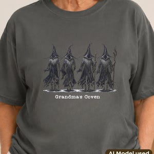 May include: A charcoal grey short-sleeve t-shirt featuring a graphic illustration of four figures in dark robes and pointed hats standing in a row. Below the figures, the text "Grandma's Coven" is printed in a white, slightly distressed font.