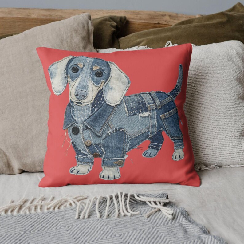 May include: A square cushion with a dachshund dog illustration. The dog is wearing a denim jacket and trousers, set against a coral background. The dog's ears and paws are light blue. The cushion is a home decor item.