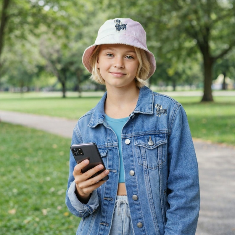 May include: A light pink bucket hat and denim jacket with a dachshund dog design. The hat and jacket are worn by a person holding a smartphone. The background is a park with green grass and trees.