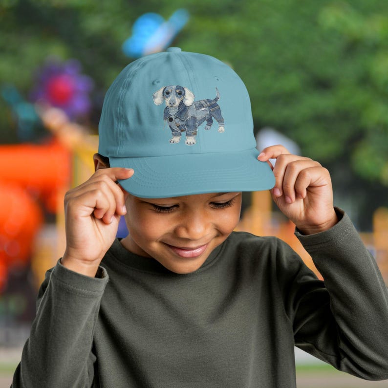 May include: A light blue baseball cap with a dachshund dog illustration. The dog is wearing a denim jacket. The cap has a curved peak and a classic six-panel design, suitable for casual wear.