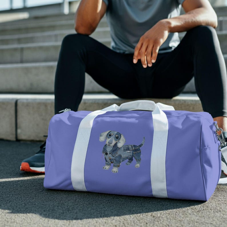 May include: A lilac duffel bag with white straps and a zip. The bag has a watercolour illustration of a dachshund wearing a denim jacket and trousers. The bag is designed for carrying sports or gym gear.