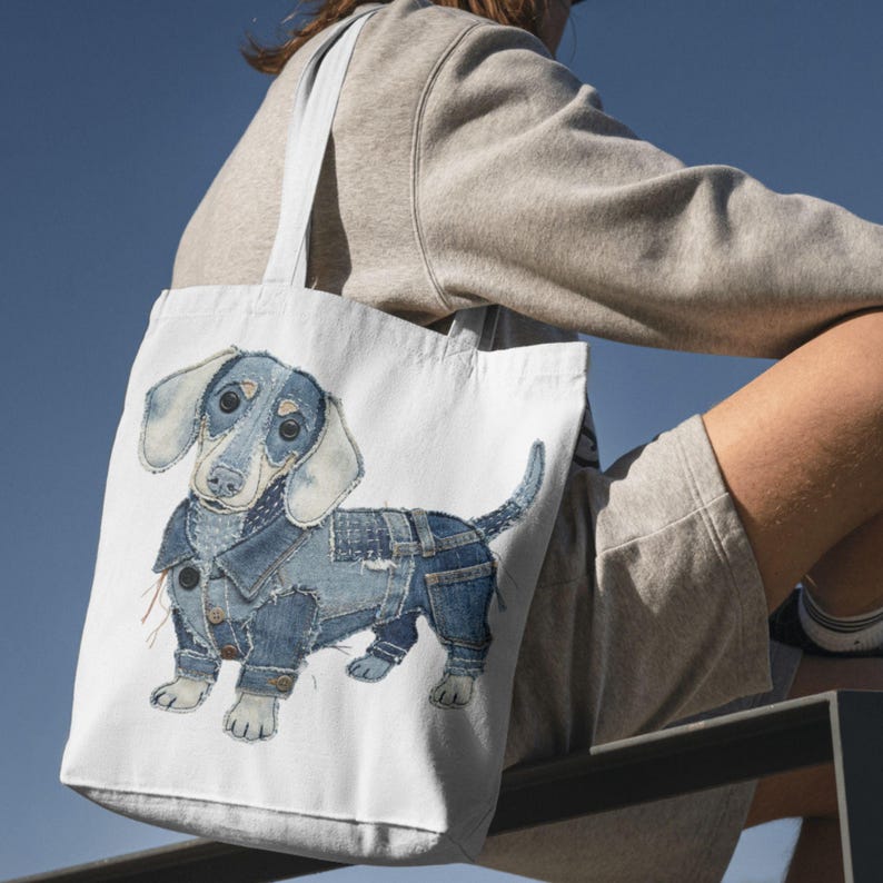 May include: White tote bag with a watercolour illustration of a dachshund in denim clothing. The dog has blue and white fur. The bag has two white handles. The background is white.