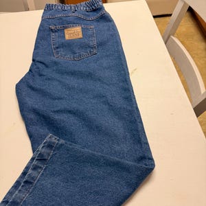 May include: Medium blue denim jeans featuring an elasticized waistband and a rectangular tan patch on the back pocket with the text Casual Joe JEANS. The jeans have a classic five-pocket design with visible stitching and a straight leg cut.