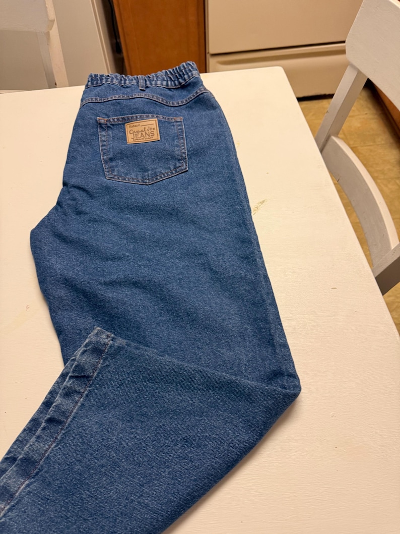 May include: Medium blue denim jeans featuring an elasticized waistband and a rectangular tan patch on the back pocket with the text Casual Joe JEANS. The jeans have a classic five-pocket design with visible stitching and a straight leg cut.