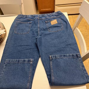 May include: Blue denim jeans featuring an elastic waistband, two back pockets, and a tan rectangular patch on the right back pocket with the text Camel Joe JEANS. The jeans have a straight-leg cut with stitched hems at the bottom of each leg.