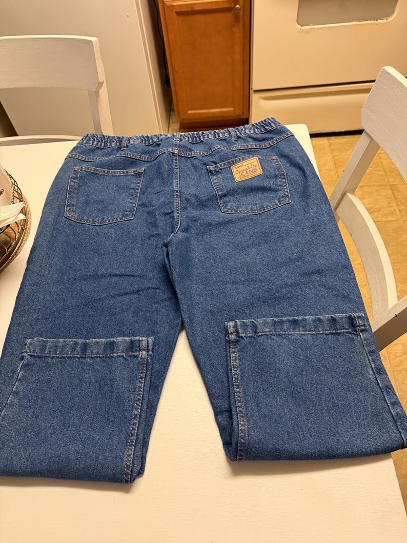 May include: Blue denim jeans featuring an elastic waistband, two back pockets, and a tan rectangular patch on the right back pocket with the text Camel Joe JEANS. The jeans have a straight-leg cut with stitched hems at the bottom of each leg.