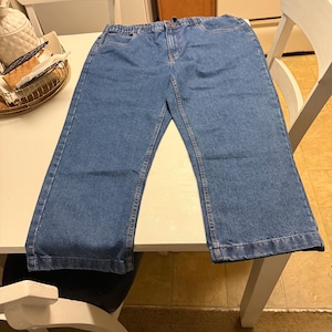 May include: A pair of medium-wash blue denim jeans featuring an elastic waistband, a front zipper fly with a button closure, two front pockets, and straight-cut legs with stitched hems. The denim fabric has a classic textured appearance.