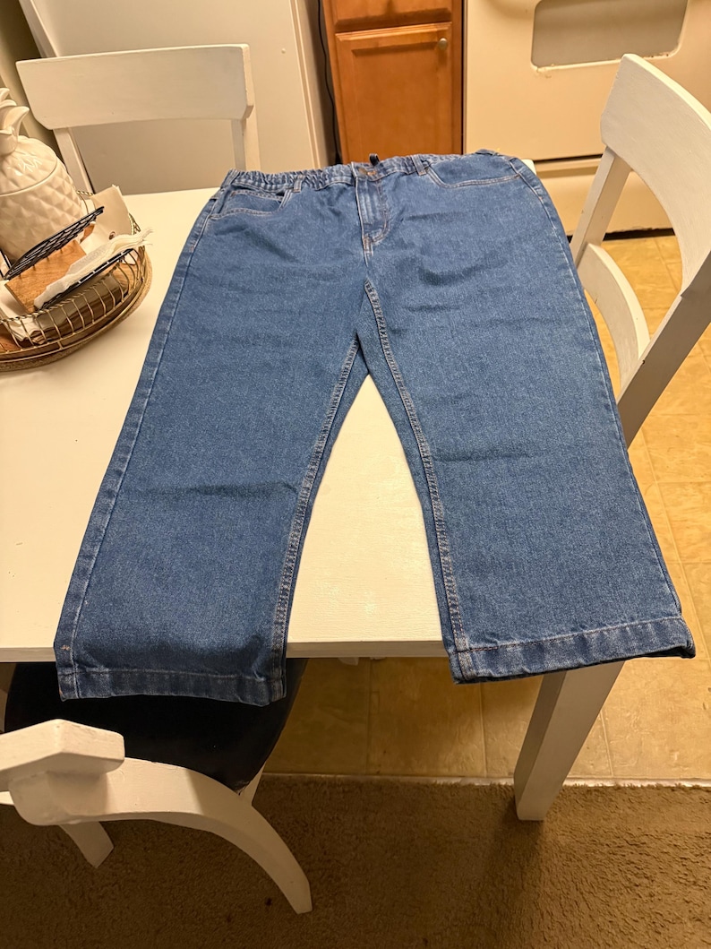 May include: A pair of medium-wash blue denim jeans featuring an elastic waistband, a front zipper fly with a button closure, two front pockets, and straight-cut legs with stitched hems. The denim fabric has a classic textured appearance.
