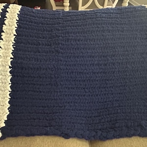 Könnte beinhalten: Eine marineblaue, handgestrickte Decke mit zwei vertikalen Streifen in Weiß und Grau. Die Decke ist aus dickem Garn gefertigt und hat ein strukturiertes Aussehen, geeignet für ein Bett oder Sofa. Die Decke ist über einem Sofa gefaltet.