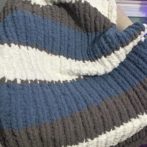 Puede incluir: Una manta de punto grueso con un diseño de rayas. La manta presenta amplias rayas blancas, azules y grises. El material suave y texturizado está doblado, mostrando la naturaleza acogedora y hecha a mano de la manta.