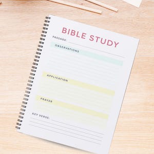 Op de afbeelding: Een wit notitieboek met spiraalbinding en de tekst "Bible Study" in roze letters. Het notitieboek heeft secties voor "Passage", "Observations", "Application", "Prayer" en "Key Verse".