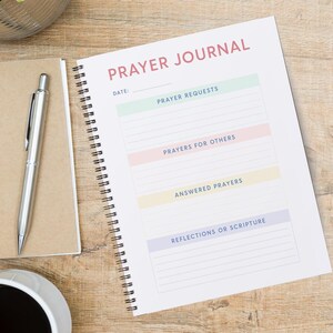 May include: A white prayer journal with a spiral binding. The cover has the text "PRAYER JOURNAL" in pink letters. The journal has sections for prayer requests, prayers for others, answered prayers, and reflections or scripture.
