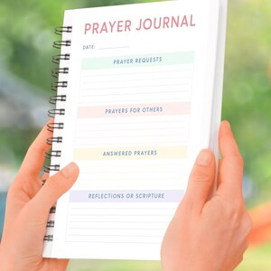 May include: A white prayer journal with a black spiral binding. The cover has the words "PRAYER JOURNAL" in pink lettering. The journal has sections for "Prayer Requests", "Prayers for Others", "Answered Prayers", and "Reflections or Scripture".