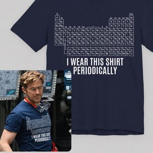 May include: A navy blue short-sleeved t-shirt featuring a white graphic of the periodic table of elements printed on the chest. Below the table, the text reads I WEAR THIS SHIRT PERIODICALLY in white capital letters. The shirt has a crew neckline.
