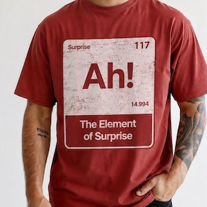 Könnte beinhalten: Ein ziegelrotes Kurzarm-T-Shirt mit einer weißen rechteckigen Grafik, die an ein Element des Periodensystems erinnert. Der Text lautet Surprise, 117, Ah!, 14.994 und The Element of Surprise. Die Grafik hat eine abgenutzte Textur.