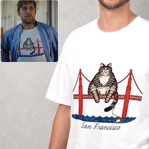 May include: A white short-sleeved t-shirt featuring a graphic illustration of a brown tabby cat sitting on the Golden Gate Bridge. Below the bridge, the text San Francisco is printed in a cursive black font. The cat holds a fishing line.