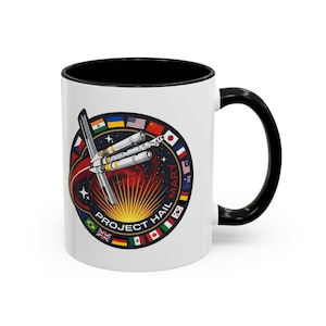 Peut inclure: Un mug en céramique blanc avec un intérieur et une anse noirs, orné d'un motif circulaire représentant un vaisseau spatial dans l'espace. Le design inclut le texte PROJECT HAIL MARY et une bordure de divers drapeaux internationaux.