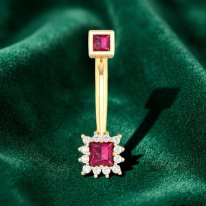 May include: A gold belly button ring featuring a square-cut red gemstone at the top and a larger square-cut red gemstone at the bottom, surrounded by a halo of small, clear, round-cut white stones in a prong setting on a curved gold barbell.