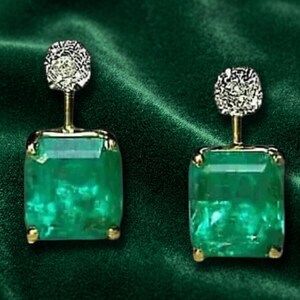 Puede incluir: Un par de pendientes colgantes con grandes piedras preciosas verdes de corte rectangular engastadas en metal dorado. Cada pendiente tiene un poste superior circular texturizado conectado a la piedra verde mediante un fino eje vertical.