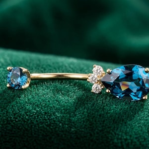Pode incluir: Uma joia corporal de barra curva dourada com uma pedra preciosa azul-escura de corte redondo numa extremidade e uma pedra azul-escura em forma de pera acentuada por três pequenas pedras redondas transparentes na outra, em metal dourado polido.