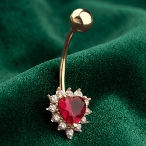 May include: A gold-toned curved barbell belly ring featuring a central heart-shaped red stone surrounded by a halo of small clear white stones. The piece includes a smooth gold-toned ball at the top end and a decorative heart-shaped charm at the base.