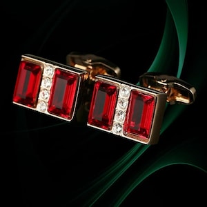 May include: A pair of gold-toned cufflinks with rectangular, ruby-red gemstones. Each cufflink features two large red stones and a row of smaller, clear stones. The background is black with a green, flowing design.