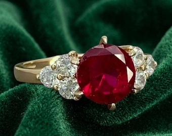 May include: A gold ring featuring a large, round, deep red centre stone flanked by three smaller, clear, round-cut stones on each side. The band is polished gold, and the setting holds the vibrant red gemstone securely in a prong-style arrangement.