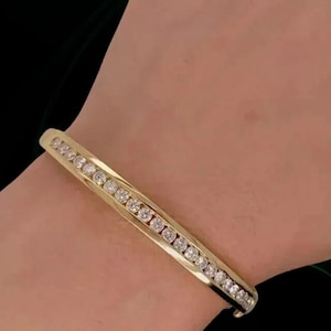 Può includere: Un braccialetto color oro con una fila di pietre preziose rotonde e trasparenti. Il braccialetto è un braccialetto a cerniera e le pietre preziose sono molto vicine. Il braccialetto è indossato su un polso.