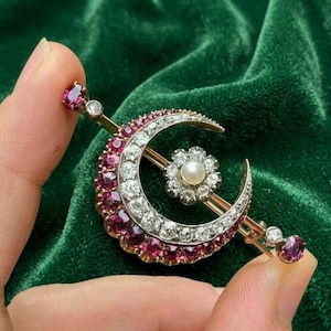 May include: A crescent moon brooch featuring a row of round-cut red rubies and clear white diamonds. A central flower-shaped cluster sits within the crescent, set with a single white pearl surrounded by small diamonds, all mounted on a gold bar.