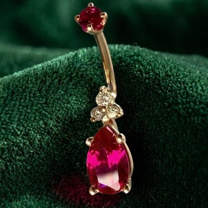 May include: A gold-toned navel ring featuring a curved barbell with a small round red stone at the top. The bottom dangle component consists of three small clear stones arranged in a cluster above a larger, pear-shaped red stone in a prong setting.